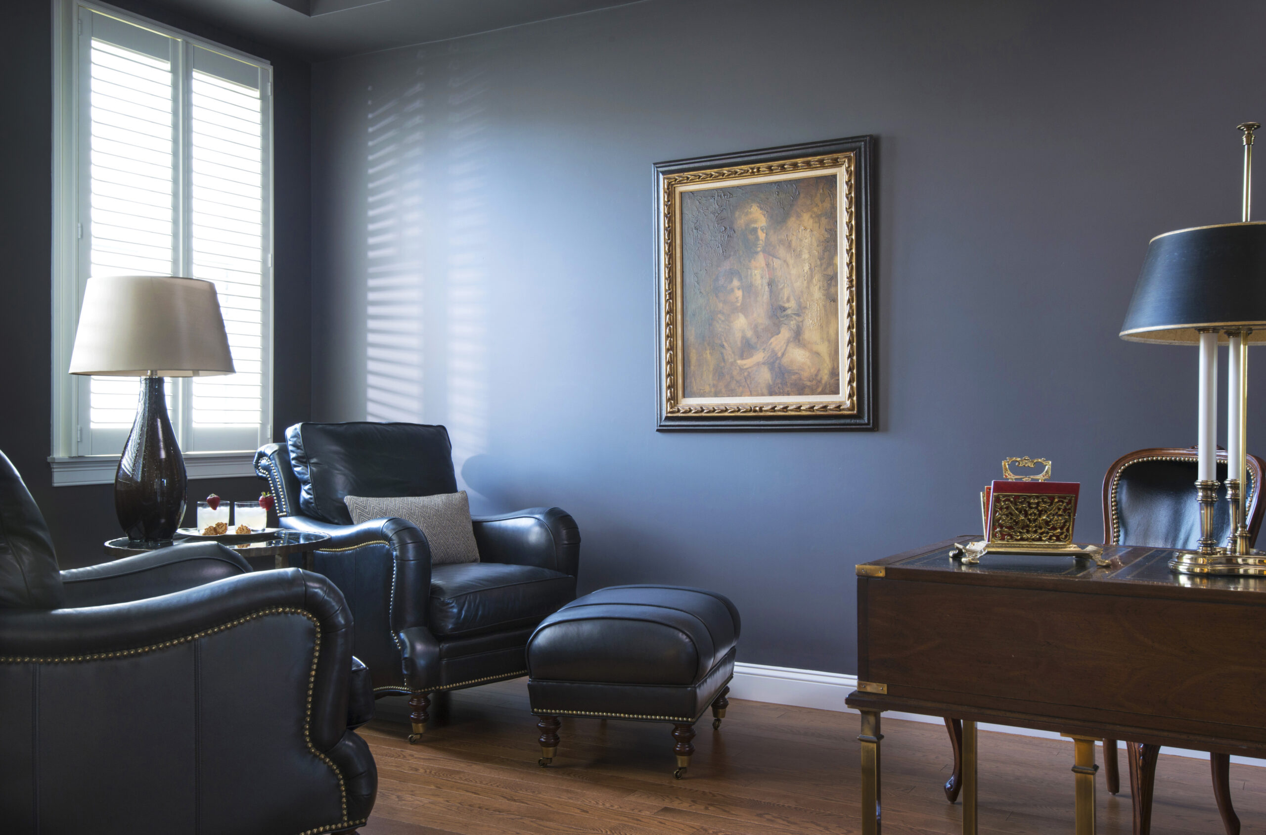 A traditional study with dark leather armchairs, a wooden desk, a framed painting on a blue wall, and a window with shutters letting in light—perfectly showcasing luxury interior design by an expert Interior Designer in Massachusetts.