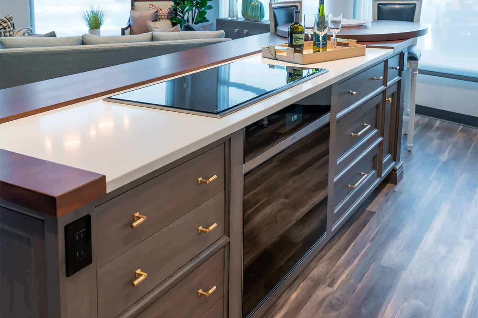 Modern kitchen island with an electric cooktop, gray cabinetry with gold handles, and a tray with wine and glasses on a white countertop—an example of luxury interior design by a top Interior Designer Massachusetts.