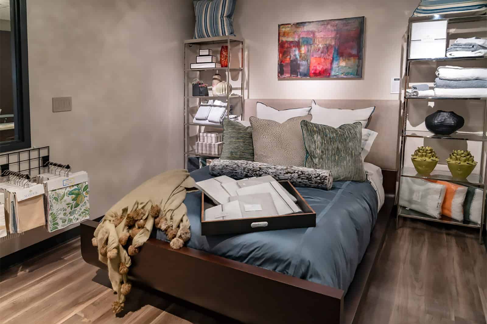 A neatly made bed with multiple pillows and a tray sits in a modern bedroom, curated with Luxury Interior Design touches, surrounded by shelves with folded linens and decorative items on a wooden floor.