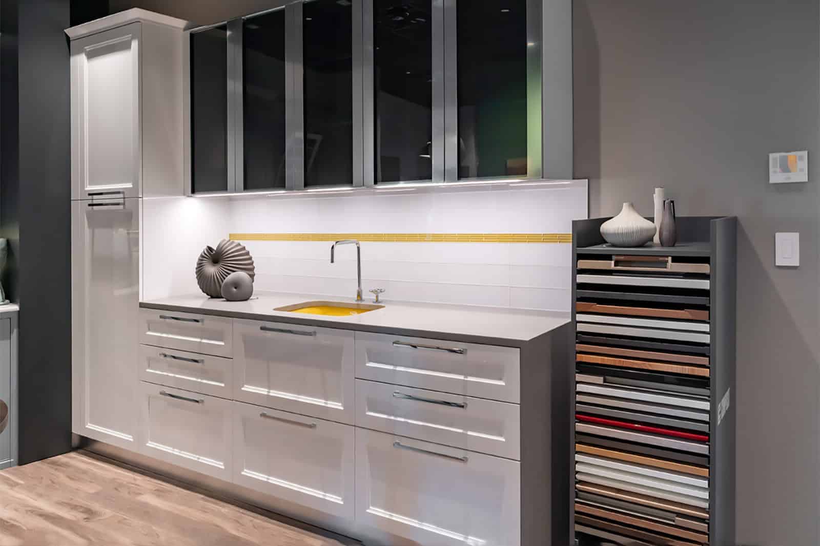 Modern kitchen with glossy white cabinets, a small sink, decorative vases, and a display rack holding countertop samples against a gray wall—perfectly styled by an expert Interior Designer Massachusetts for luxury interior design enthusiasts.