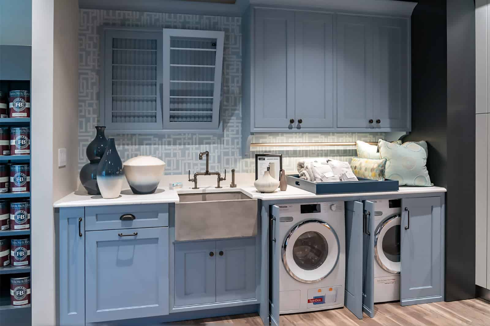 A modern laundry room designed with luxury interior design touches features light blue cabinets, a farmhouse sink, washer and dryer, decorative vases, shelves, and a pull-out ironing board—perfect inspiration from an interior designer Massachusetts.