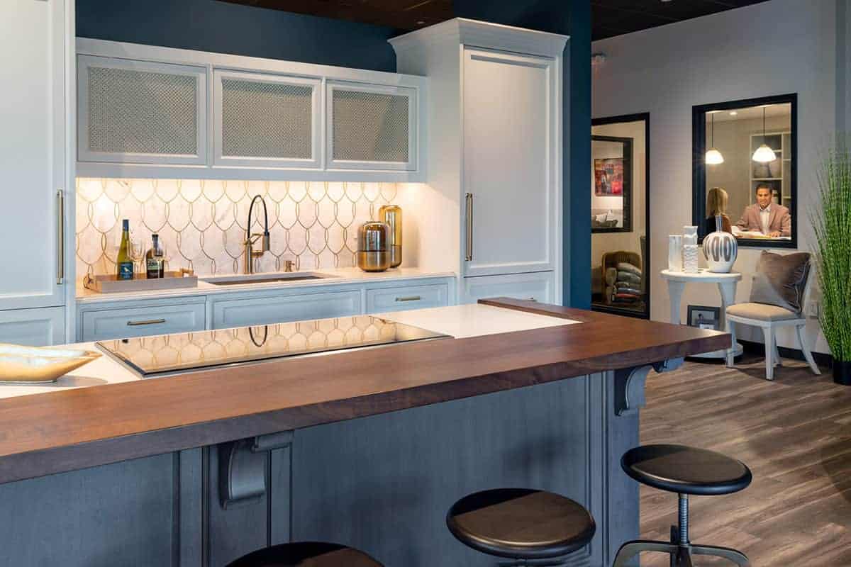 Modern kitchen with white cabinets, wooden countertop, bar stools, built-in appliances, and geometric tile backsplash; crafted with Luxury Interior Design expertise. Two people are visible in an adjacent room through a window.
