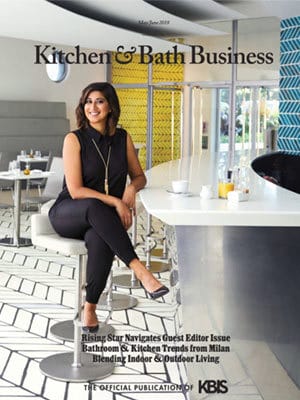 A woman in black attire sits on a modern white barstool in a contemporary kitchen with patterned walls, showcasing luxury interior design on the cover of Kitchen & Bath Business magazine.