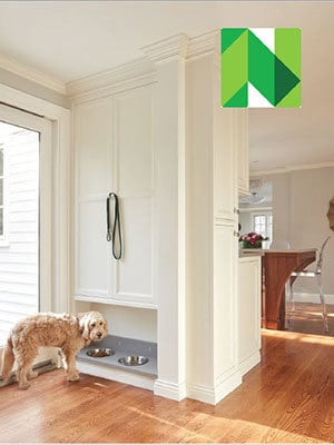 A dog stands by a built-in pet feeding station with two bowls and a leash hanging on the wall in a bright, white room with hardwood floors, showcasing luxury interior design by an interior designer from Massachusetts.