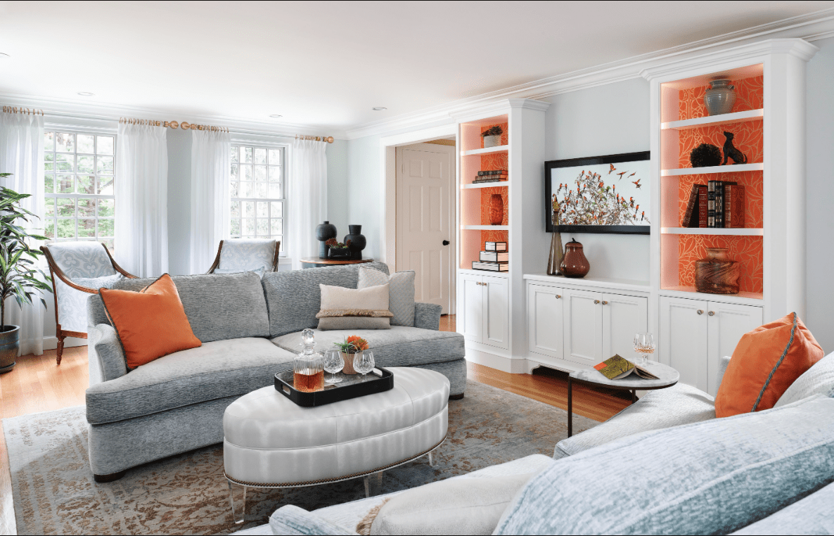 Bright living room with light blue sectional sofa, orange accent pillows, built-in white shelves with orange backing, large window, and a white ottoman serving as a coffee table, a welcoming space brought to life through full service interior design.