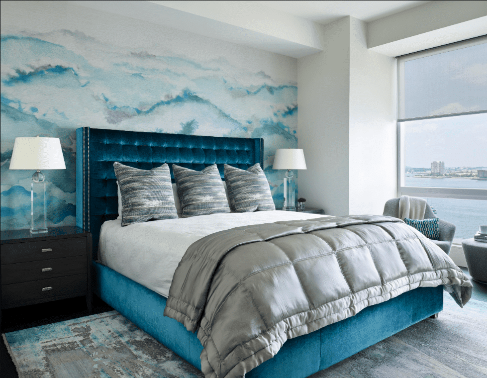 Modern bedroom expertly crafted through full service interior design, featuring a teal upholstered bed, gray bedding, abstract blue wall art, two bedside tables with lamps, and a large window overlooking a stunning cityscape.
