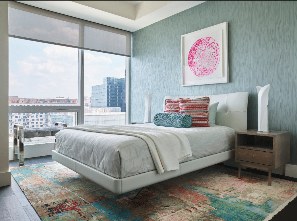 Modern bedroom with a white bed, colorful pillows, abstract rug, nightstands, large window, and wall art above the bed.