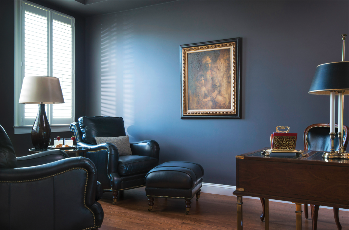 A study with dark leather chairs, a wooden desk with lamps, a framed painting on a blue wall, hardwood floor, and light from a window with white shutters.