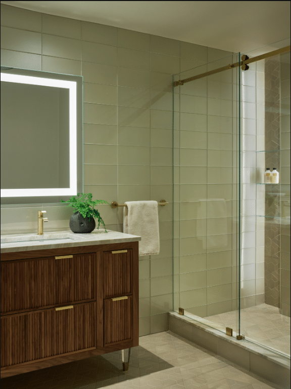 Modern bathroom with a lighted mirror, wood vanity with gold fixtures, towel on a gold bar, and glass-enclosed shower. Small plant decorates the vanity.