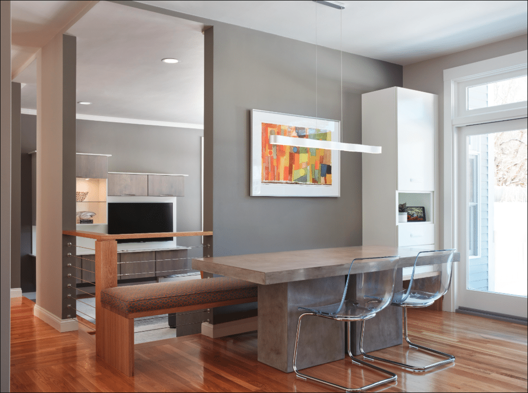 Modern dining area with a concrete table, bench seating, two clear chairs, abstract wall art, and large windows letting in natural light.