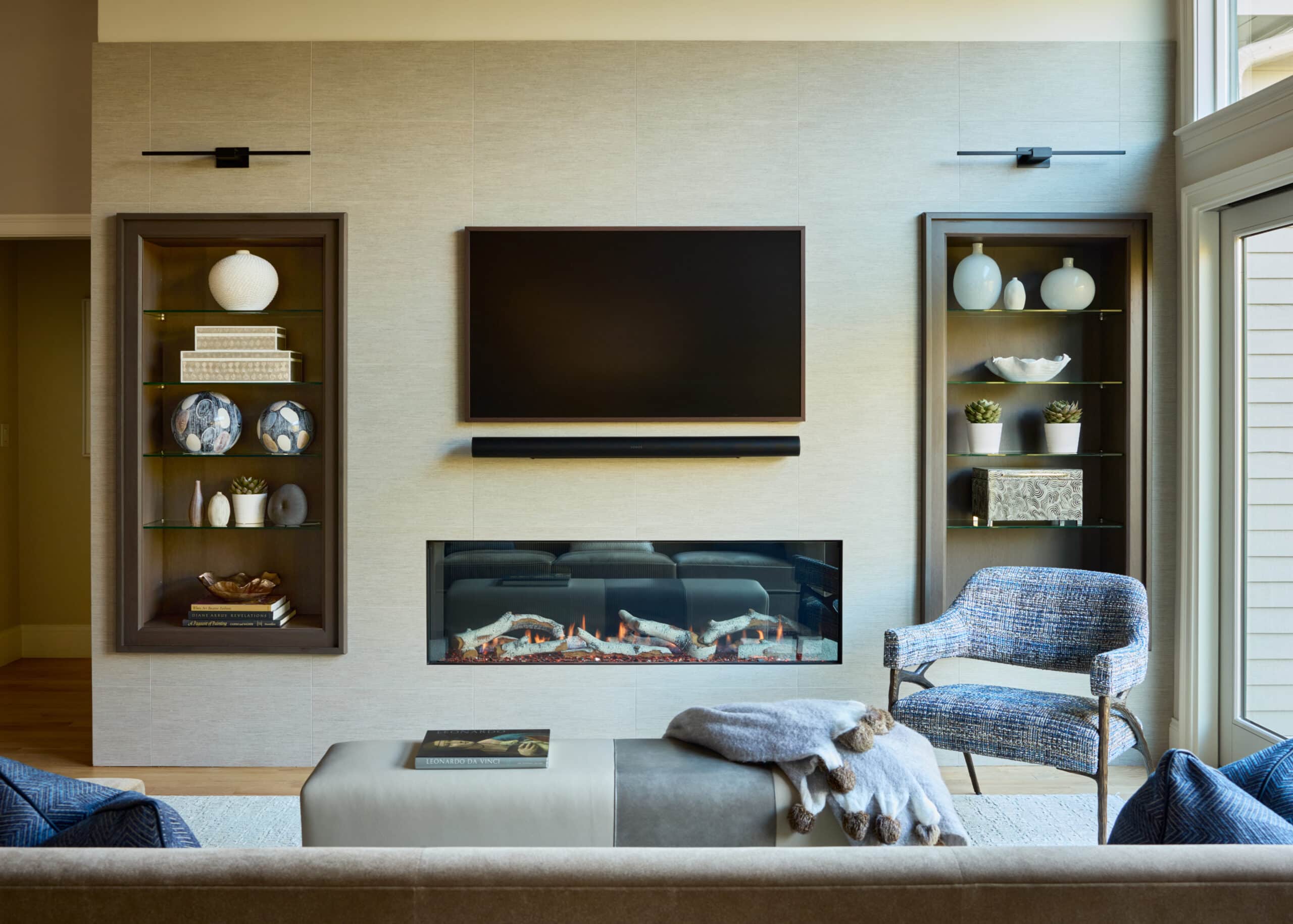 Modern living room interior design featuring a wall-mounted TV and fireplace, flanked by built-in shelves displaying decor items; blue armchair and ottoman with throw blanket in the foreground create a cozy yet stylish atmosphere.